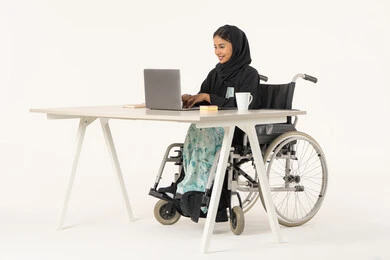 تعليم ذوي الهمم استخدام الأجهزة التقنية المتطورة، جودة حياة ذوي الهمم في الامارات, بورتريه لامرأة عربية اماراتية خليجية محجبة مبتسمة من ذوي الاحتياجات الخاصة ترتدي العباءة السوداء و تجلس على كرسي متحرك و تقوم بالنقر على لوحة المفاتيح  عبر جهاز اللابتوب, خلفية بيضاء