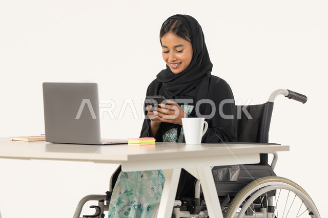 دعم المجتمع الاماراتي لذوي الهمم، العمل عن بعد عبر الجوال، بورتريه لامرأة عربية اماراتية خليجية محجبة مبتسمة من ذوي الاحتياجات الخاصة ترتدي العباءة السوداء و تجلس على كرسي متحرك أمام جهاز الكمبيوتر و تقوم بالعمل على الهاتف المحمول، خلفية بيضاء