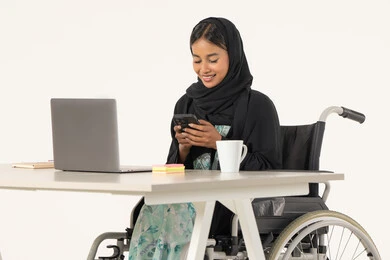 Supporting the UAE community for people with disabilities, working remotely via mobile, portrait of a smiling veiled Emirati Gulf Arab woman with special needs wearing a black abaya, sitting in a wheelchair in front of a computer and working on a mobile phone, white background