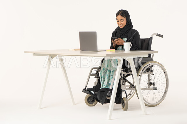 الإمساك بالهاتف المحمول وتصفح مواقع الانترنت و معرفة آخر الأخبار والمستجدات، بورتريه لامرأة عربية اماراتية خليجية محجبة مبتسمة من ذوي الاحتياجات الخاصة ترتدي العباءة السوداء و تجلس على كرسي متحرك أمام جهاز الكمبيوتر و تقوم بالعمل على الموبايل، خلفية بيضاء