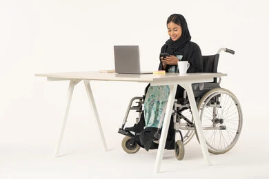 الإمساك بالهاتف المحمول وتصفح مواقع الانترنت و معرفة آخر الأخبار والمستجدات، بورتريه لامرأة عربية اماراتية خليجية محجبة مبتسمة من ذوي الاحتياجات الخاصة ترتدي العباءة السوداء و تجلس على كرسي متحرك أمام جهاز الكمبيوتر و تقوم بالعمل على الموبايل، خلفية بيضاء