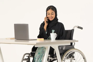 انجاز صفقات الاعمال عن طريق الموبايل، التواصل مع العملاء عبر الجوال،  بورتريه لامرأة عربية اماراتية خليجية محجبة مبتسمة من ذوي الاحتياجات الخاصة ترتدي العباءة السوداء و تجلس على كرسي متحرك أمام جهاز الكمبيوتر و تقوم بالعمل على الهاتف المحمول، خلفية بيضاء