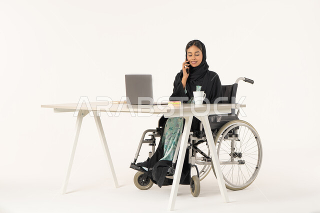 Answering and responding to phone calls via mobile phone, portrait of a smiling veiled Emirati Gulf Arab woman with special needs wearing a black abaya and sitting on a wheelchair in front of a computer and talking on a mobile phone, white background