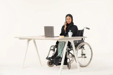الرد والاجابة على الاتصالات الهاتفية عبر الهاتف المحمول،  بورتريه لامرأة عربية اماراتية خليجية محجبة مبتسمة من ذوي الاحتياجات الخاصة ترتدي العباءة السوداء و تجلس على كرسي متحرك أمام جهاز الكمبيوتر و تقوم بالتحدث من خلال الموبايل، خلفية بيضاء