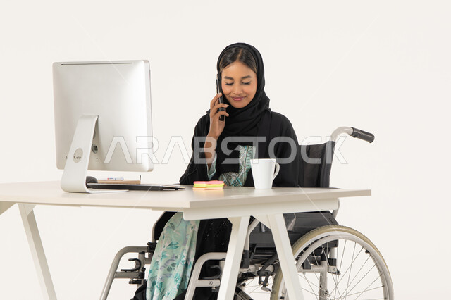 اجراء مكالمة هاتفية، العمل عن بعد عبر الجوال، بورتريه لامرأة عربية اماراتية خليجية محجبة مبتسمة من ذوي الاحتياجات الخاصة ترتدي العباءة السوداء و تجلس على كرسي متحرك أمام جهاز الكمبيوتر و تقوم بالعمل على الهاتف المحمول، خلفية بيضاء