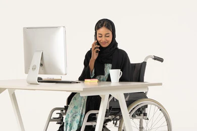Making a phone call, working remotely via mobile, portrait of a smiling veiled Emirati Gulf Arab woman with special needs wearing a black abaya and sitting on a wheelchair in front of a computer and working on a mobile phone, white background