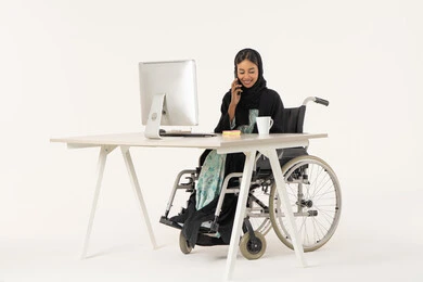 التواصل مع العملاء عبر الجوال، الرد والاجابة على الاتصالات الهاتفية عبر الهاتف المحمول، بورتريه لامرأة عربية اماراتية خليجية محجبة مبتسمة من ذوي الاحتياجات الخاصة ترتدي العباءة السوداء و تجلس على كرسي متحرك أمام جهاز الكمبيوتر و تقوم بالتحدث من خلال الموبايل، خلفية بيضاء