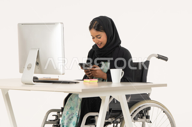 العمل عن بعد عبر الجوال، بورتريه لامرأة عربية اماراتية خليجية محجبة مبتسمة من ذوي الاحتياجات الخاصة ترتدي العباءة السوداء و تجلس على كرسي متحرك أمام جهاز الكمبيوتر و تقوم بالعمل على الهاتف المحمول، خلفية بيضاء