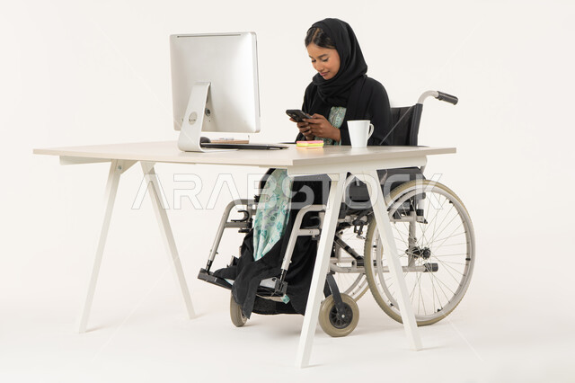 تصفح مواقع أخبار السوشال ميديا, العمل عن بعد عبر الجوال، بورتريه لامرأة عربية اماراتية خليجية محجبة مبتسمة من ذوي الاحتياجات الخاصة ترتدي العباءة السوداء و تجلس على كرسي متحرك أمام جهاز الكمبيوتر و تقوم بالعمل على الهاتف المحمول، خلفية بيضاء