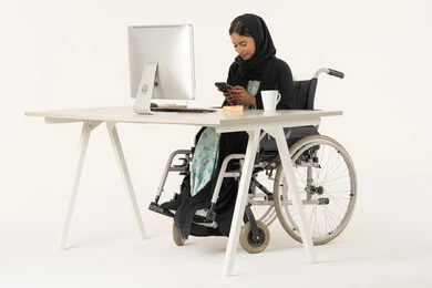 تصفح مواقع أخبار السوشال ميديا, العمل عن بعد عبر الجوال، بورتريه لامرأة عربية اماراتية خليجية محجبة مبتسمة من ذوي الاحتياجات الخاصة ترتدي العباءة السوداء و تجلس على كرسي متحرك أمام جهاز الكمبيوتر و تقوم بالعمل على الهاتف المحمول، خلفية بيضاء