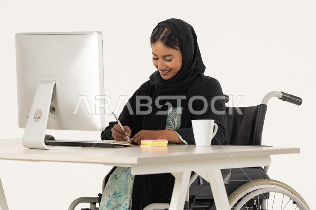 اعداد التقارير و الملفات الخاصة بالعمل، ايماءات التركيز و التمعن, بورتريه لامرأة عربية اماراتية خليجية محجبة مبتسمة من ذوي الاحتياجات الخاصة ترتدي العباءة السوداء و تجلس على كرسي متحرك أمام جهاز الكمبيوتر و تمسك بيدها قلم تقوم بتدوين الملاحظات على اوراق، خلفية بيضاء