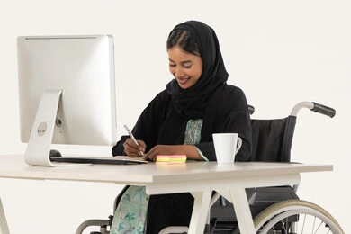 اعداد التقارير و الملفات الخاصة بالعمل، ايماءات التركيز و التمعن, بورتريه لامرأة عربية اماراتية خليجية محجبة مبتسمة من ذوي الاحتياجات الخاصة ترتدي العباءة السوداء و تجلس على كرسي متحرك أمام جهاز الكمبيوتر و تمسك بيدها قلم تقوم بتدوين الملاحظات على اوراق، خلفية بيضاء