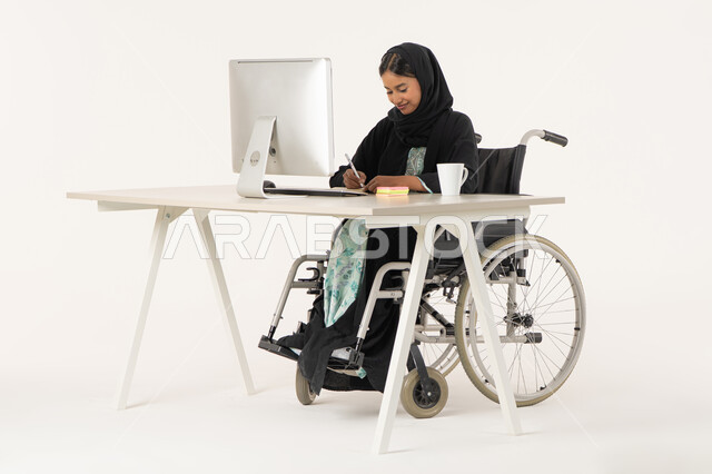ايماءات التركيز و التمعن, بورتريه لامرأة عربية اماراتية خليجية محجبة من ذوي الاحتياجات الخاصة ترتدي العباءة السوداء و تجلس على كرسي متحرك أمام جهاز الكمبيوتر و تمسك بيدها قلم تقوم بتدوين الملاحظات على اوراق، اعداد التقارير و الملفات الخاصة بالعمل، خلفية بيضاء