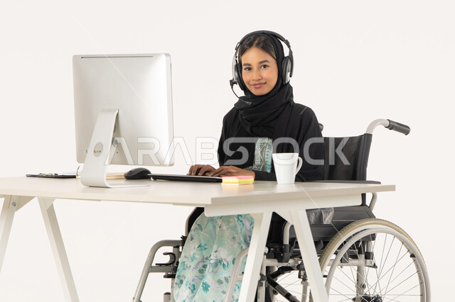 الجلوس وراء المكتب وتنفيذ الأعمال على الأجهزة الحديثة,  بورتريه لامرأة عربية اماراتية خليجية محجبة مبتسمة تنظر الى الكاميرا من ذوي الاحتياجات الخاصة ترتدي العباءة السوداء و تجلس على كرسي متحرك أمام جهاز الكمبيوتر و تضع سماعات الهيدفون, مهن ووظائف ادارية مكتبية, إدارة شؤون الأعمال و تنظيمها، خلفية بيضاء