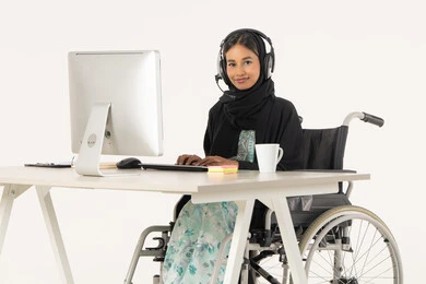 الجلوس وراء المكتب وتنفيذ الأعمال على الأجهزة الحديثة,  بورتريه لامرأة عربية اماراتية خليجية محجبة مبتسمة تنظر الى الكاميرا من ذوي الاحتياجات الخاصة ترتدي العباءة السوداء و تجلس على كرسي متحرك أمام جهاز الكمبيوتر و تضع سماعات الهيدفون, مهن ووظائف ادارية مكتبية, إدارة شؤون الأعمال و تنظيمها، خلفية بيضاء
