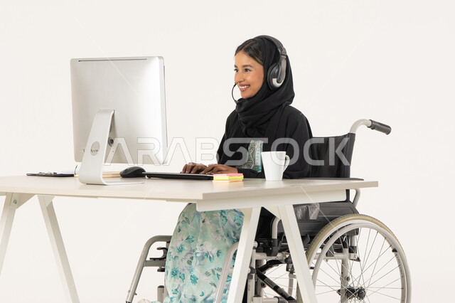 الإجابة على أسئلة العملاء، بورتريه لامرأة عربية اماراتية خليجية محجبة من ذوي الاحتياجات الخاصة ترتدي العباءة السوداء و تجلس على كرسي متحرك أمام جهاز الكمبيوتر و تضع سماعات الهيدفون, مهن ووظائف ادارية مكتبية, إدارة شؤون الأعمال و تنظيمها، خلفية بيضاء