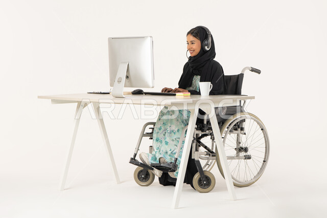 مفهوم الاستعلامات والاستفسارات، العمل في مجال خدمة العملاء، بورتريه لامرأة عربية اماراتية خليجية محجبة من ذوي الاحتياجات الخاصة ترتدي العباءة السوداء و تجلس على كرسي متحرك أمام جهاز الكمبيوتر و تضع سماعات الهيدفون, مهن ووظائف ادارية مكتبية، خلفية بيضاء