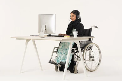 Working in customer service, portrait of a veiled Emirati Gulf Arab woman with special needs wearing a black abaya and sitting in a wheelchair in front of a computer and wearing headphones, office administrative professions and jobs, business affairs management and organization, white background