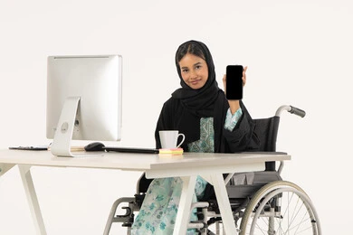Blank black screen, use of modern technologies, quality of mobile phone applications and services, portrait of a veiled Emirati Gulf Arab woman with special needs wearing a black abaya and sitting on a wheelchair in front of a computer and looking at the camera with a smile, white background