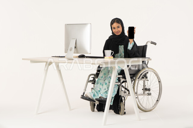 Using modern technology, displaying a blank black screen, quality of mobile phone applications and services, portrait of a veiled Arab Emirati Gulf woman with special needs wearing a black abaya and sitting in a wheelchair in front of a computer and looking at the camera with a smile, white background