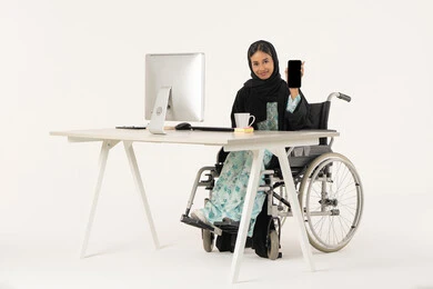 استخدام التقنيات الحديثة، عرض شاشة سوداء فارغة، جودة تطبيقات و خدمات الهاتف المحمول، بورتريه لامرأة عربية اماراتية خليجية محجبة من ذوي الاحتياجات الخاصة ترتدي العباءة السوداء و تجلس على كرسي متحرك أمام جهاز الكمبيوتر و تنظر للكاميرا بابتسامة, خلفية بيضاء