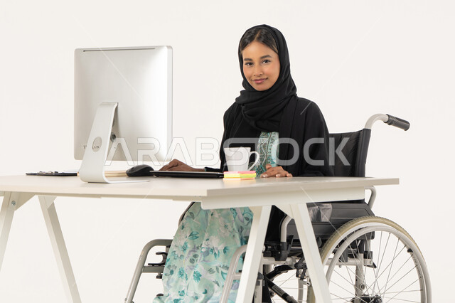 النظر للكاميرا بايماءات السعادة, مهن ووظائف ادارية مكتبية, إدارة شؤون الأعمال وتنظيمها، بورتريه لامرأة عربية اماراتية خليجية محجبة من ذوي الاحتياجات الخاصة ترتدي العباءة السوداء و تجلس على كرسي متحرك و تباشر عملها من خلال الكمبيوتر و بجانبها مشروب دافئ, خلفية بيضاء