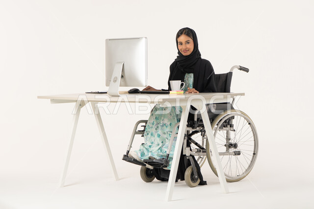النظر إلى الكاميرا بتعابير الثقة بالنفس، مهن ووظائف ادارية مكتبية, إدارة شؤون الأعمال وتنظيمها، بورتريه لامرأة عربية اماراتية خليجية محجبة مبتسمة من ذوي الاحتياجات الخاصة ترتدي العباءة السوداء و تجلس على كرسي متحرك و تباشر عملها من خلال الكمبيوتر و بجانبها مشروب دافئ, خلفية بيضاء