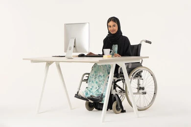 النظر إلى الكاميرا بتعابير الثقة بالنفس، مهن ووظائف ادارية مكتبية, إدارة شؤون الأعمال وتنظيمها، بورتريه لامرأة عربية اماراتية خليجية محجبة مبتسمة من ذوي الاحتياجات الخاصة ترتدي العباءة السوداء و تجلس على كرسي متحرك و تباشر عملها من خلال الكمبيوتر و بجانبها مشروب دافئ, خلفية بيضاء