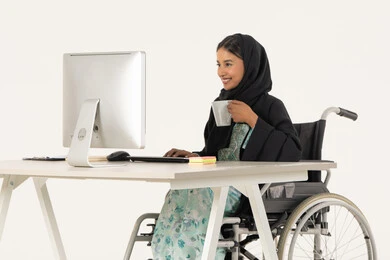 الحيوية والنشاط عند شرب الكافيين، مهن ووظائف ادارية مكتبية, إدارة شؤون الأعمال وتنظيمها، بورتريه لامرأة عربية اماراتية خليجية محجبة من ذوي الاحتياجات الخاصة ترتدي العباءة السوداء و تجلس على كرسي متحرك و تباشر عملها من خلال الكمبيوتر و تشرب مشروب دافئ, خلفية بيضاء