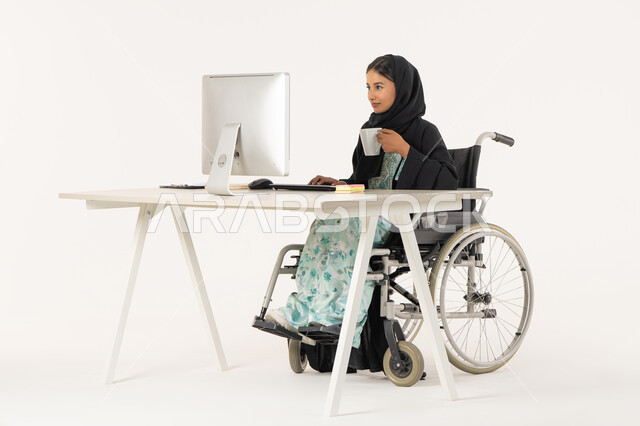 الاستمتاع بتناول المشروبات, العمل لتحقيق الاهداف و الاستراتيجيات، مهن ووظائف ادارية مكتبية, إدارة شؤون الأعمال وتنظيمها، بورتريه لامرأة عربية اماراتية خليجية محجبة من ذوي الاحتياجات الخاصة ترتدي العباءة السوداء و تجلس على كرسي متحرك و تباشر عملها من خلال الكمبيوتر و تشرب مشروب دافئ, خلفية بيضاء