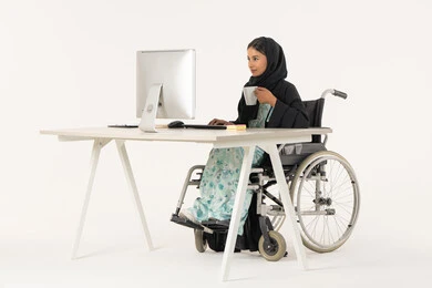 Enjoying drinks, working to achieve goals and strategies, office administrative professions and jobs, business management and organization, portrait of a veiled Arab Emirati Gulf woman with special needs wearing a black abaya, sitting in a wheelchair, working on the computer and drinking a warm drink, white background