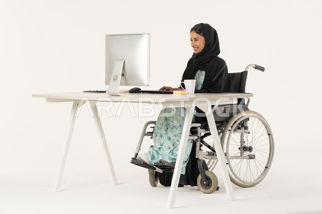 العمل لتحقيق الاهداف و الاستراتيجيات، مهن ووظائف ادارية مكتبية, إدارة شؤون الأعمال وتنظيمها،  بورتريه لامرأة عربية اماراتية خليجية محجبة من ذوي الاحتياجات الخاصة ترتدي العباءة السوداء و تجلس على كرسي متحرك و تباشر عملها من خلال الكمبيوتر, خلفية بيضاء