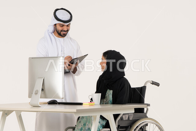 Writing and noting something, discussing work between colleagues, office administrative professions and jobs, portrait of a veiled Arab Emirati Gulf woman with special needs wearing a black abaya and sitting in a wheelchair and discussing with her Emirati colleague who is wearing a white kandura and ghutra and holding a paper holder, white background