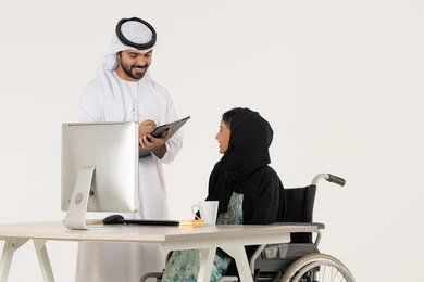 Writing and noting something, discussing work between colleagues, office administrative professions and jobs, portrait of a veiled Arab Emirati Gulf woman with special needs wearing a black abaya and sitting in a wheelchair and discussing with her Emirati colleague who is wearing a white kandura and ghutra and holding a paper holder, white background