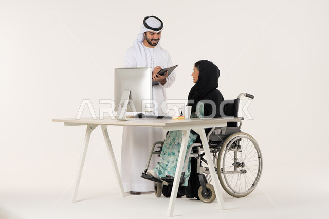Writing and recording notes, discussing work between colleagues, office administrative professions and jobs, portrait of a veiled Arab Emirati Gulf woman with special needs wearing a black abaya and sitting in a wheelchair, discussing with her Emirati colleague who is wearing a white kandura and ghutra and holding a paper holder, white background