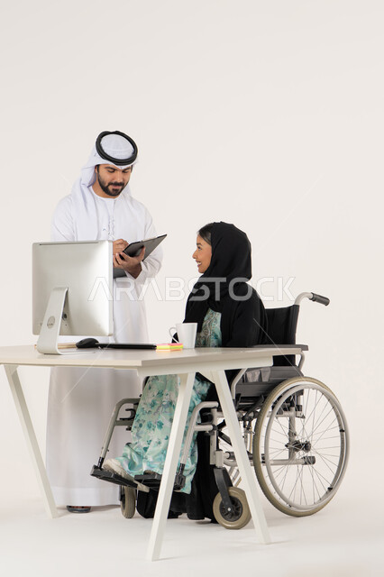 التدوين و الكتابة على الورق, مناقشة عمل بين الزملاء, مهن ووظائف ادارية مكتبية, بورتريه لامرأة عربية اماراتية خليجية محجبة من ذوي الاحتياجات الخاصة ترتدي العباءة السوداء و تجلس على كرسي متحرك و تتناقش مع زميلها الاماراتي الذي برتدي الكندورة والغترة البيضاء و يمسك حامل الأوراق, خلفية بيضاء