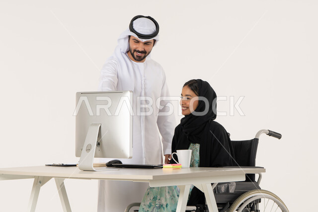 الاشارة الى شيء ما, العمل لتحقيق الاهداف والاستراتيجيات, بورتريه لامرأة عربية اماراتية خليجية محجبة من ذوي الاحتياجات الخاصة ترتدي العباءة السوداء و تجلس على كرسي متحرك تستخدم الحاسب المحمول و تتناقش مع زميلها الذي برتدي الكندورة والغترة البيضاء, خلفية بيضاء