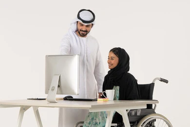 الاشارة الى شيء ما, العمل لتحقيق الاهداف والاستراتيجيات, بورتريه لامرأة عربية اماراتية خليجية محجبة من ذوي الاحتياجات الخاصة ترتدي العباءة السوداء و تجلس على كرسي متحرك تستخدم الحاسب المحمول و تتناقش مع زميلها الذي برتدي الكندورة والغترة البيضاء, خلفية بيضاء