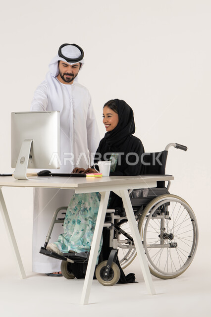 Study and analysis of projects, pointing to something, working to achieve goals and strategies, portrait of a veiled Emirati Gulf Arab woman with special needs wearing a black abaya and sitting in a wheelchair using a laptop and discussing with her colleague who is wearing a white kandura and ghutra, white background