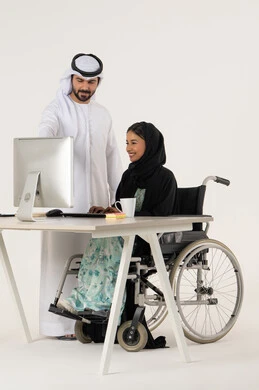 Study and analysis of projects, pointing to something, working to achieve goals and strategies, portrait of a veiled Emirati Gulf Arab woman with special needs wearing a black abaya and sitting in a wheelchair using a laptop and discussing with her colleague who is wearing a white kandura and ghutra, white background