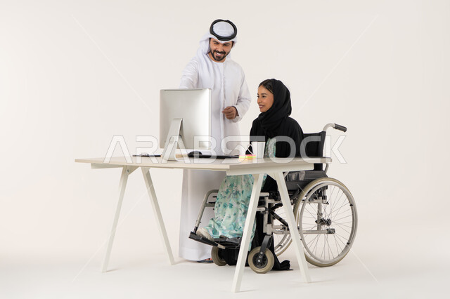 Unity and teamwork, pointing to something, working to achieve goals and strategies, portrait of a veiled Emirati Gulf Arab woman with special needs wearing a black abaya and sitting in a wheelchair using a laptop and discussing with her colleague wearing a white kandura and ghutra, white background