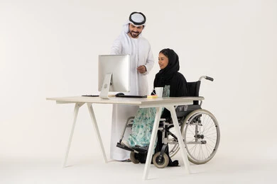 الوحدة و العمل الجماعي, الاشارة الى شيء ما, العمل لتحقيق الاهداف والاستراتيجيات, بورتريه لامرأة عربية اماراتية خليجية محجبة من ذوي الاحتياجات الخاصة ترتدي العباءة السوداء و تجلس على كرسي متحرك تستخدم الحاسب المحمول و تتناقش مع زميلها الذي برتدي الكندورة والغترة البيضاء, خلفية بيضاء