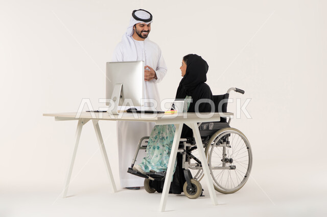 مناقشة عمل بين الزملاء, مهن ووظائف ادارية مكتبية, بورتريه لامرأة عربية اماراتية خليجية محجبة من ذوي الاحتياجات الخاصة ترتدي العباءة السوداء و تجلس على كرسي متحرك و تتناقش مع زميلها الاماراتي المبتسم الذي برتدي الكندورة والغترة البيضاء, خلفية بيضاء