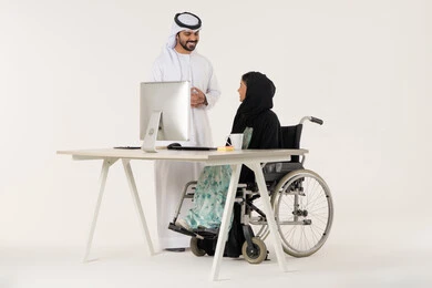 مناقشة عمل بين الزملاء, مهن ووظائف ادارية مكتبية, بورتريه لامرأة عربية اماراتية خليجية محجبة من ذوي الاحتياجات الخاصة ترتدي العباءة السوداء و تجلس على كرسي متحرك و تتناقش مع زميلها الاماراتي المبتسم الذي برتدي الكندورة والغترة البيضاء, خلفية بيضاء