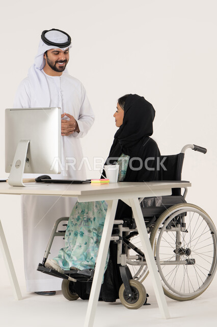 أهمية النقاش و الحوار في العمل, مهن ووظائف ادارية مكتبية, بورتريه لامرأة عربية اماراتية خليجية محجبة من ذوي الاحتياجات الخاصة ترتدي العباءة السوداء و تجلس على كرسي متحرك و تتناقش مع زميلها الاماراتي المبتسم الذي برتدي الكندورة والغترة البيضاء, خلفية بيضاء
