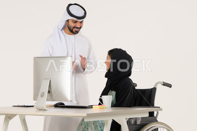 تحقيق اهداف الشركه, بيئة العمل الاماراتي, الوحده و العمل الجماعي, مهن ووظائف ادارية مكتبية, بورتريه لامرأة عربية اماراتية خليجية محجبة من ذوي الاحتياجات الخاصة ترتدي العباءة السوداء و تجلس على كرسي متحرك و تتناقش مع زميلها الذي برتدي الكندورة والغترة البيضاء, خلفية بيضاء