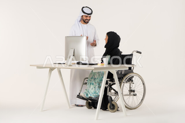 تفاعل إيجابي في بيئة العمل, تأمين معدات للمساعده على الحركة والتنقل, العمل لتحقيق الاهداف والاستراتيجيات، مهن ووظائف ادارية مكتبية, بورتريه لامرأة عربية اماراتية خليجية محجبة من ذوي الاحتياجات الخاصة ترتدي العباءة السوداء و تجلس على كرسي متحرك و تتناقش مع زميلها الذي برتدي الكندورة والغترة البيضاء, خلفية بيضاء