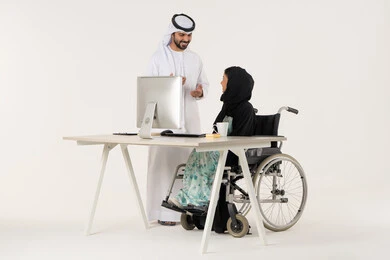 Positive interaction in the work environment, providing equipment to assist mobility, working to achieve goals and strategies, office administrative professions and jobs, portrait of a veiled Arab Emirati Gulf woman with special needs wearing a black abaya and sitting in a wheelchair, discussing with her colleague who is wearing a white kandura and ghutra, white background