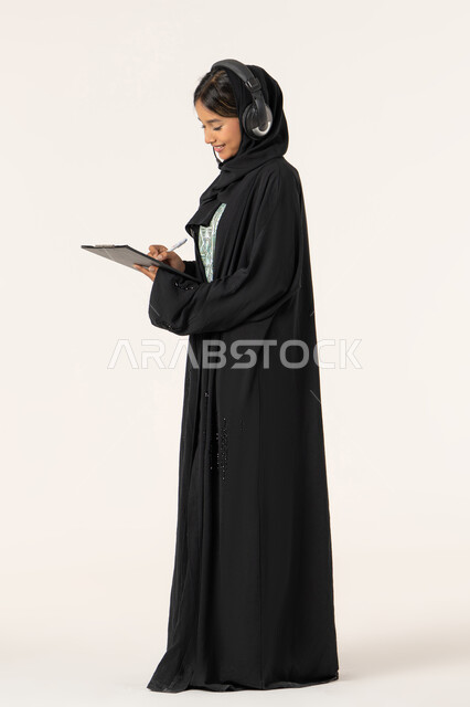 مفهوم خدمة العملاء، متابعة وتطوير العمل، تدوين و تسجيل الملاحظات و الشكاوي، بورتريه من الجانب لموظفة عربية خليجية اماراتية محجبة مبتسمة ترتدي عباءة سوداء تضع سماعات الهيدفون على رأسها و تقوم بالكتابة على الاوراق، الرد على الاستفسارت والتساؤولات، خلفية بيضاء