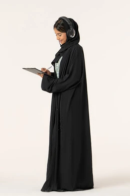 مفهوم خدمة العملاء، متابعة وتطوير العمل، تدوين و تسجيل الملاحظات و الشكاوي، بورتريه من الجانب لموظفة عربية خليجية اماراتية محجبة مبتسمة ترتدي عباءة سوداء تضع سماعات الهيدفون على رأسها و تقوم بالكتابة على الاوراق، الرد على الاستفسارت والتساؤولات، خلفية بيضاء